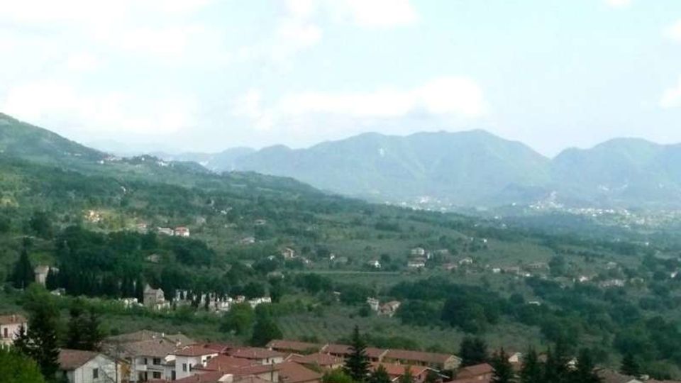 Veduta panoramica di un paese con case dai tetti in terracotta in primo piano, circondato da campi e boschi. Sullo sfondo si estendono colline e montagne sotto un cielo parzialmente nuvoloso.