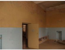 Interno di una stanza spoglia con pareti superiori intonacate color ocra e fascia inferiore rivestita di piastrelle azzurre, con segni di deterioramento e umidità. Al centro c’è una porta aperta che conduce a un ambiente più buio, con pavimento in piastrelle e una finestra con grata visibile sullo sfondo.