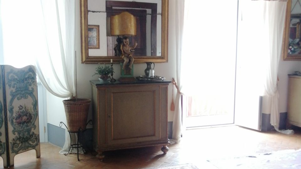 Salotto in stile classico con pavimento in parquet e grande tappeto rosso, credenza con specchio dorato e decorazioni, e soffitto con fregi ornamentali. Ampie porte-finestre con tende bianche e mantovane fiorate illuminano la stanza.