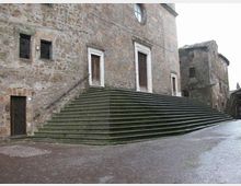 Facciata in pietra di un edificio storico con ampie scalinate in pietra verde che salgono lungo l’angolo fino a un ingresso con cornice chiara. Davanti si apre una piazza pavimentata in mattoni, con altri edifici antichi sullo sfondo.