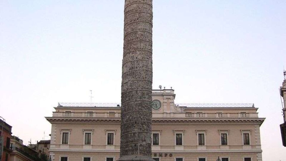 Alta colonna monumentale in pietra con bassorilievi a spirale e statua in cima, situata al centro di una piazza urbana. Sullo sfondo si vede un grande edificio neoclassico con portici ad archi, facciata simmetrica e un orologio centrale.