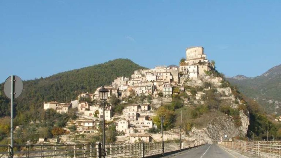 Strada asfaltata su un ponte con ringhiere metalliche che conduce verso un borgo di case in pietra arroccato su una collina. In cima al paese si distingue una grande torre o edificio fortificato, con montagne boscose sullo sfondo sotto un cielo sereno.