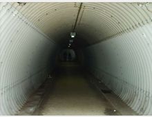 Tunnel pedonale sotterraneo con pareti e soffitto semicircolari in pannelli metallici ondulati, illuminato da una fila di lampade a soffitto. Il pavimento in cemento corre dritto verso un’uscita luminosa in fondo al passaggio.