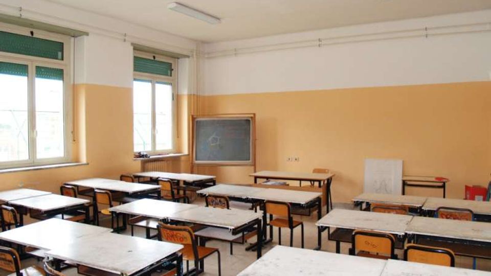 Aula scolastica con file di banchi e sedie rivolti verso una lavagna, pareti bicolori e illuminazione a neon sul soffitto. Grandi finestre laterali con tapparelle verdi e un tavolo dell’insegnante lungo la parete di fondo.