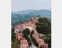 Veduta panoramica di un borgo collinare con case dai tetti in coppi rossi e una strada centrale alberata che sale verso una chiesa con campanile. Sullo sfondo si vedono colline e montagne verdi con abitazioni sparse nella valle.