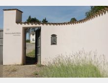 Muro d’ingresso intonacato bianco con copertura in coppi, un cancello nero socchiuso e una cassetta postale in metallo fissata al centro. Attraverso l’apertura si vede un vialetto che conduce a un edificio bianco sullo sfondo, con vegetazione e cielo sereno intorno.