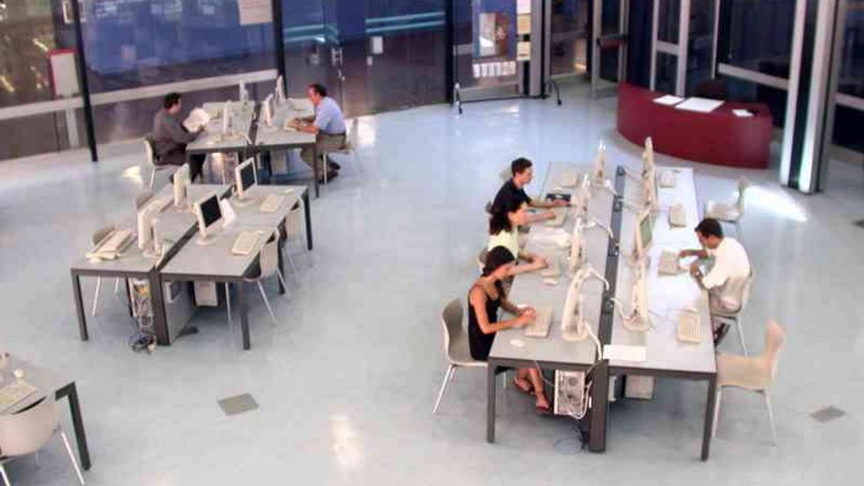 Ampio spazio interno tipo laboratorio informatico con pavimento chiaro, tavoli disposti in isole e numerosi computer desktop. Sullo sfondo ci sono pareti vetrate, porte d’ingresso e un bancone curvo vicino all’entrata.
