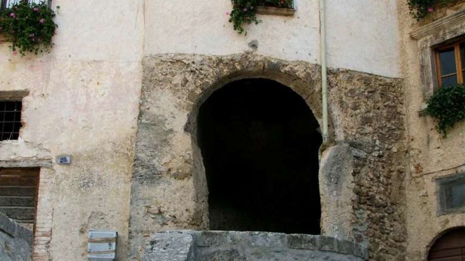 Facciata di un edificio storico in pietra e intonaco con un grande arco d’ingresso che conduce a un passaggio coperto. In primo piano si vedono una scalinata e un muretto in pietra, con finestre e piccoli balconi con ringhiere in ferro e fioriere.