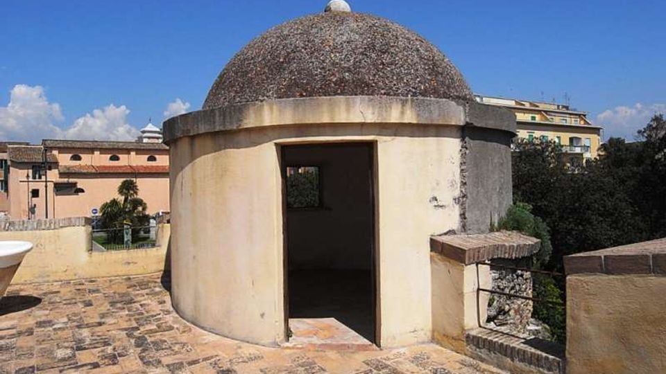 Piccolo edificio circolare in muratura con cupola scura e ingresso aperto, situato su una terrazza pavimentata in pietra. Sullo sfondo si vedono edifici cittadini, alberi e cielo sereno.