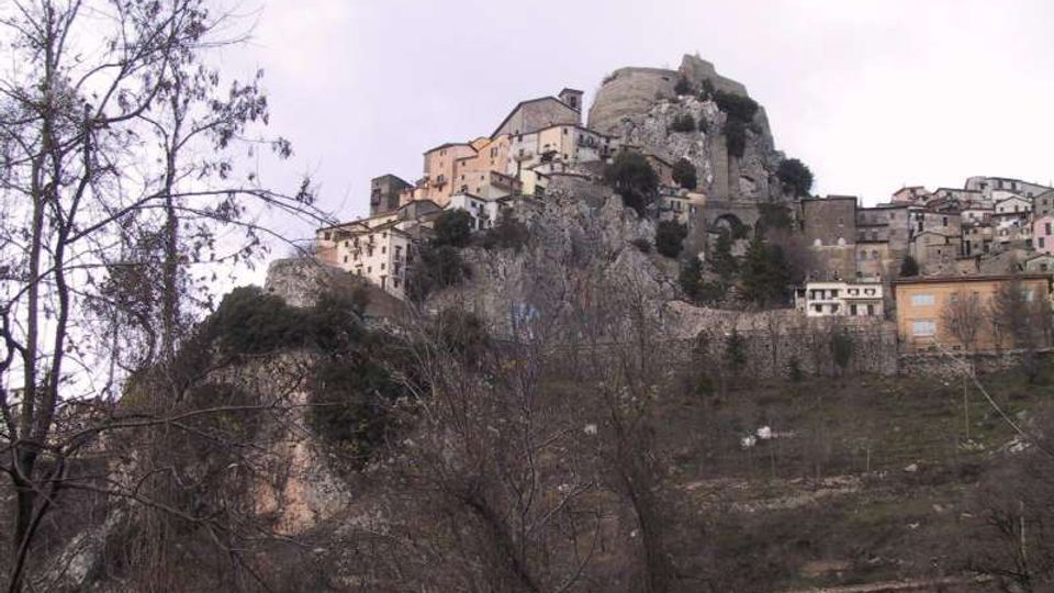 Borgo storico arroccato su una rupe rocciosa, con case in pietra addossate al pendio e resti di una fortificazione in cima. In primo piano si vedono alberi spogli e terreni collinari sotto un cielo nuvoloso.