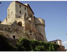 Castello in pietra su uno sperone roccioso, con alte mura fortificate, archi e una torre cilindrica merlata. In primo piano vegetazione rampicante sulle pareti e cielo sereno sullo sfondo.