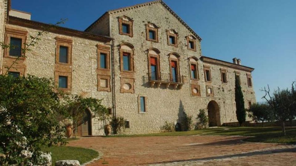 Facciata di un grande edificio storico in pietra e mattoni, con finestre incorniciate, archi e un piccolo balcone centrale. Davanti si apre un cortile con pavimentazione in mattoni, aiuole e alberi in un’area verde.