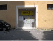Ingresso di un edificio con grande portone grigio incassato in una facciata color ocra, con un’insegna gialla “Amnesty International” sopra la porta e alcuni avvisi affissi. Davanti c’è un’area asfaltata con ombre marcate e una finestra con grata visibile sulla parete.