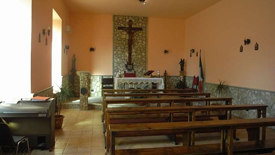 Interno di una piccola cappella con pareti color pesca e rivestimento in pietra nella zona dell’altare, con crocifisso centrale sopra l’altare. File di panche in legno su pavimento piastrellato, luce laterale da una finestra e alcune decorazioni religiose alle pareti.