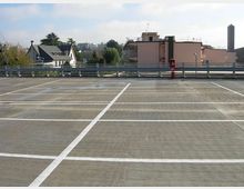 Parcheggio multipiano all’aperto con pavimentazione in cemento e linee bianche che delimitano gli stalli. Sullo sfondo si vedono ringhiere metalliche, edifici residenziali e alberi sotto un cielo parzialmente nuvoloso.