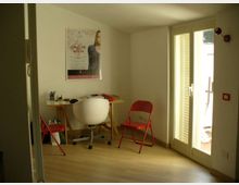 Piccolo studio con pavimento in legno chiaro, tavolo da lavoro con sedia bianca e due sedie pieghevoli rosse, e un poster appeso alla parete. Una porta-finestra con persiane lascia entrare molta luce e dà accesso a un balcone.