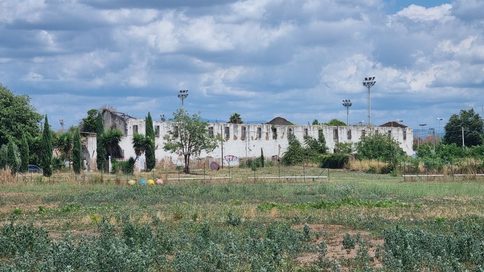 Ampio campo erboso e incolto con una recinzione bassa, alberi sparsi e alcuni pali di illuminazione. Sullo sfondo si vede un lungo edificio bianco fatiscente con mura rovinate e aperture irregolari sotto un cielo nuvoloso.