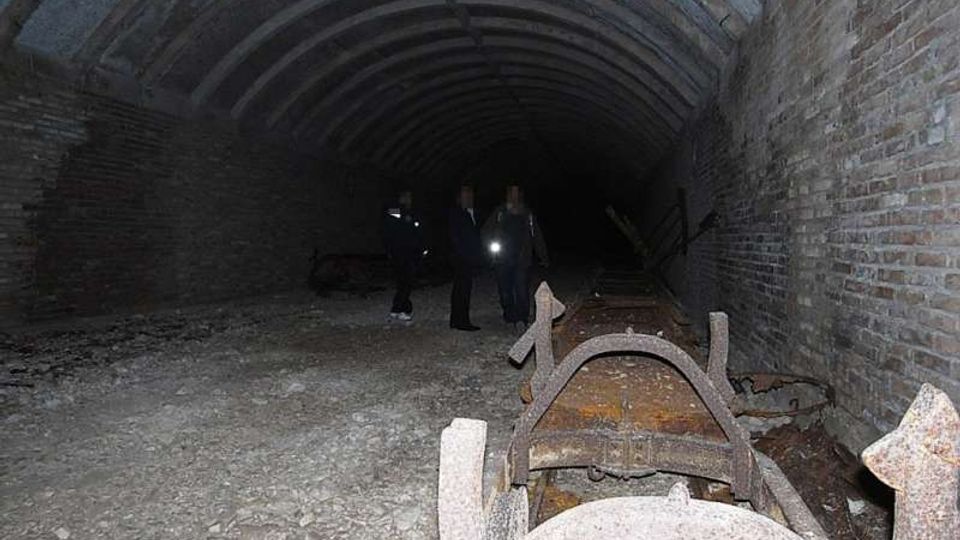 Interno di una galleria sotterranea in mattoni con soffitto a volta, pavimento in ghiaia e pareti laterali scure. In primo piano si vedono strutture metalliche arrugginite e detriti, mentre il tunnel si estende in profondità verso un’area poco illuminata.
