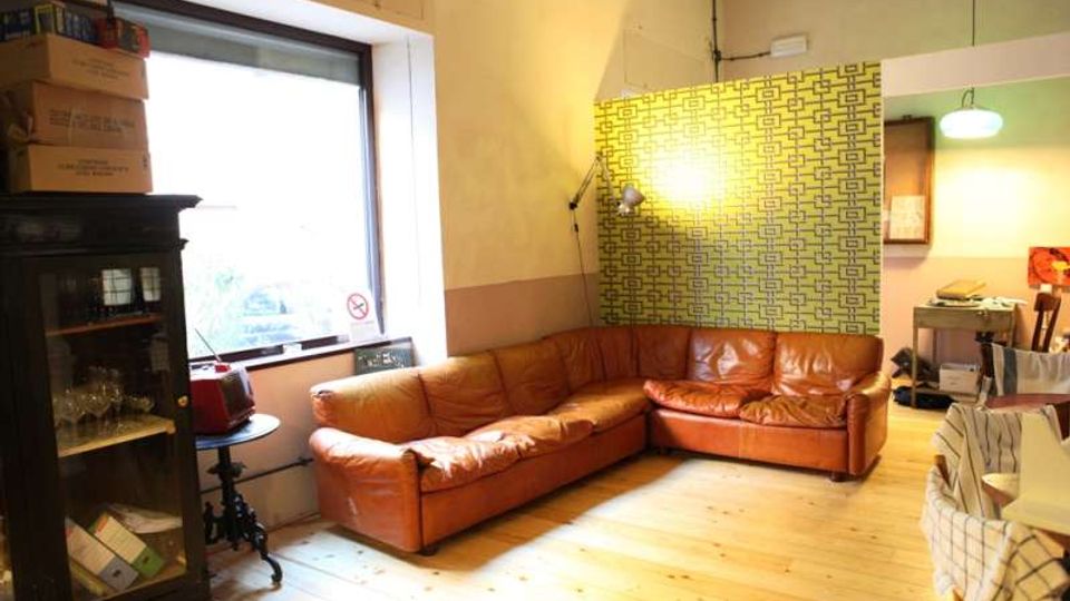 Soggiorno interno con grande divano ad angolo in pelle marrone su pavimento in legno chiaro, accanto a una finestra ampia e a una vetrina scura. Sul fondo si vede una parete con carta da parati geometrica verde e un’area adiacente con illuminazione e arredi da cucina.