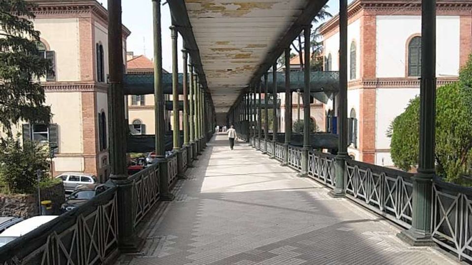 Passerella pedonale sopraelevata con lunga copertura e colonne in metallo, pavimentazione a piastrelle e ringhiere laterali. Ai lati si vedono edifici in muratura con finestre alte e tetti in tegole, con alberi e auto nel cortile sottostante.