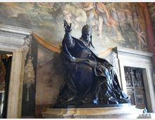 Statua monumentale in bronzo di una figura seduta su un alto basamento, collocata in un interno storico con grandi affreschi murali sullo sfondo. Ai lati si vedono portali in pietra con cornici decorative e una porta a pannelli intagliati.