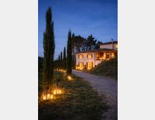 Viale di ghiaia in campagna fiancheggiato da alti cipressi, illuminato da lanterne a terra lungo il percorso. Sullo sfondo si vede una villa a due piani con persiane e luci calde accese, circondata da alberi sotto un cielo serale.