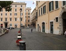 Piazza urbana con pavimentazione in sampietrini, lampioni e fioriere lungo un marciapiede, circondata da palazzi storici color ocra con finestre e persiane. Sullo sfondo si vedono facciate con cornici decorative e un’area stradale con auto parcheggiate.