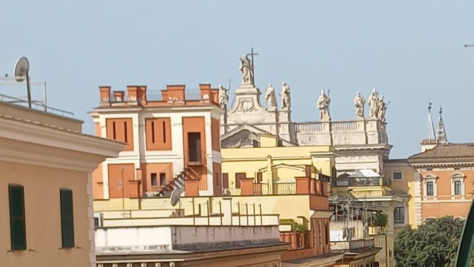 Veduta urbana con palazzi a più piani dai colori ocra e terracotta, con finestre e persiane, disposti lungo una strada. Sullo sfondo si vede un edificio monumentale con statue sul cornicione e una croce in sommità, con tetti e campanili che emergono dietro.
