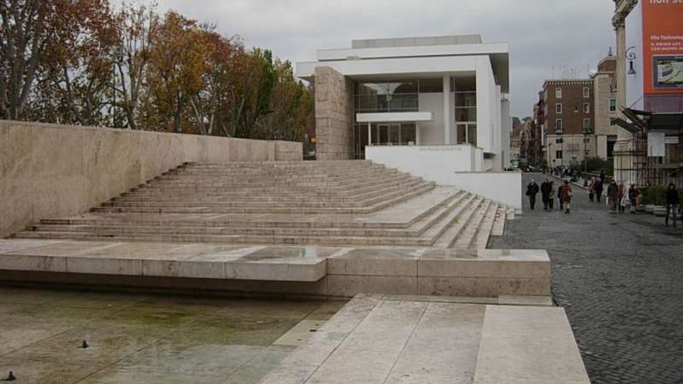 Piazza urbana con ampia scalinata in pietra chiara e vasca d’acqua in primo piano, affiancata da un viale alberato. Sullo sfondo si vede un edificio moderno bianco con grandi vetrate, con strada lastricata e palazzi lungo il lato destro.