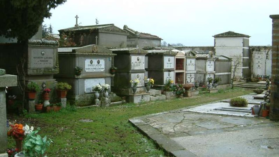 Cimitero con file di loculi e cappelle funerarie in muratura, con lapidi e vasi di fiori. Un vialetto pavimentato corre accanto a un’area erbosa, con alberi sullo sfondo.