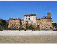 Castello costiero in pietra con due grandi bastioni cilindrici e corpo centrale rettangolare con finestre, affacciato su una spiaggia sabbiosa. Sullo sfondo si vede cielo sereno e vegetazione con palme lungo il margine della fortificazione.