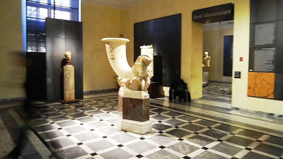 Sala museale con pavimento a scacchi in marmo e pareti gialle, con grandi pannelli espositivi neri e una finestra ad arco alta. Al centro è esposto un grande reperto scultoreo in pietra su piedistallo, con altre opere e testi informativi lungo le pareti e un passaggio verso un’altra sala.