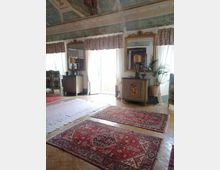 Salone interno in stile classico con soffitto decorato a affreschi e cornici, ampie finestre con tende leggere e specchi dorati alle pareti. Sul pavimento in parquet ci sono grandi tappeti orientali rossi, credenze antiche e una pianta in vaso vicino a una portafinestra.