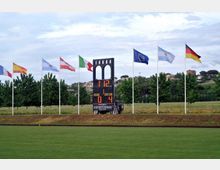 Tabellone segnapunti elettronico su un campo sportivo erboso, con una struttura centrale ad archi e cifre illuminate. Sullo sfondo una fila di alberi e diverse bandiere su alti pennoni in un’area verde collinare.