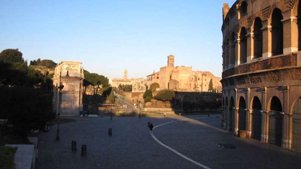 Ampia piazza lastricata accanto a un grande anfiteatro romano in mattoni con arcate sovrapposte, visto sul lato destro dell’immagine. Sullo sfondo si notano rovine e edifici storici su un lieve pendio, con alberi e lampioni lungo i bordi sotto un cielo sereno.