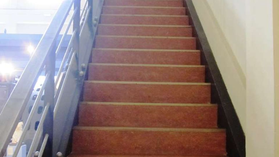 Scala interna in un edificio, con gradini in materiale rosso e corrimano metallico scuro sul lato sinistro. Pareti chiare e luce naturale proveniente dall’alto lungo il vano scala.