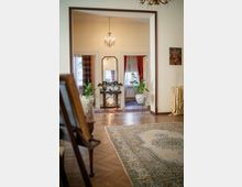 Interno di una casa in stile classico con pavimento in parquet e grande tappeto; un ampio varco incorniciato in legno conduce a un salotto con lampadario, consolle con specchio e due vasi con piante. Ai lati si vedono porte con tende rosse e a fantasia, e piccoli quadri alle pareti.