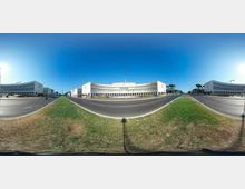 Vista panoramica a 360° di un ampio viale a più corsie con spartitraffico erboso e rotonda, circondato da grandi edifici moderni bianchi a forma curva con lunghe file di finestre. Sullo sfondo si vede un cielo sereno e alcuni alberi lungo il margine stradale.