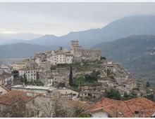 Veduta panoramica di un borgo collinare con case in pietra e tetti in coppi, disposto a terrazze attorno a un castello medievale con torre centrale. Sullo sfondo si vedono rilievi montuosi e valli boscose sotto un cielo nuvoloso.