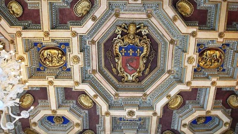Soffitto a cassettoni riccamente decorato con riquadri geometrici, inserti dorati e pannelli blu e bordeaux. Al centro spicca uno stemma araldico entro una cornice ottagonale, con un lampadario visibile sul bordo sinistro.