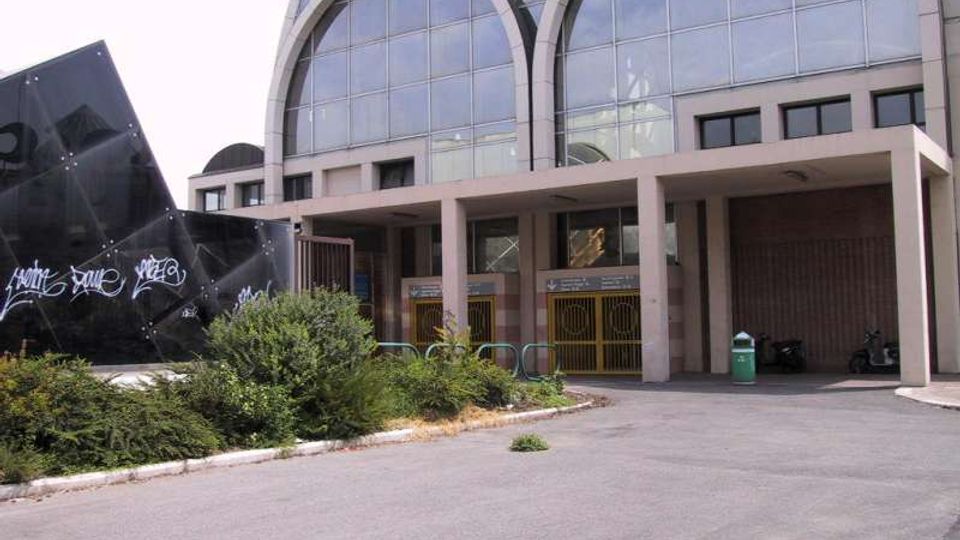 Ingresso di un edificio moderno con grande facciata vetrata ad archi e portico sorretto da pilastri, con cancelli metallici alla base. Davanti si vede un piazzale asfaltato con cespugli, una struttura nera con graffiti e un cestino verde.