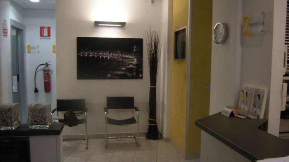 Piccola sala d’attesa interna con pavimento piastrellato e controsoffitto, due sedie metalliche, quadro a parete e illuminazione a plafoniera. A destra si vede un bancone reception, un pannello divisorio giallo con schermo integrato e un orologio a parete, con estintore e cartelli di sicurezza sul lato sinistro.