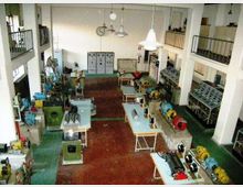 Interno di un laboratorio tecnico industriale con macchinari e motori esposti su banchi di lavoro, pavimento in mattoni rossi e pareti bianche alte. Lampade a sospensione, armadi elettrici e soppalchi con ringhiere corrono lungo i lati della sala.