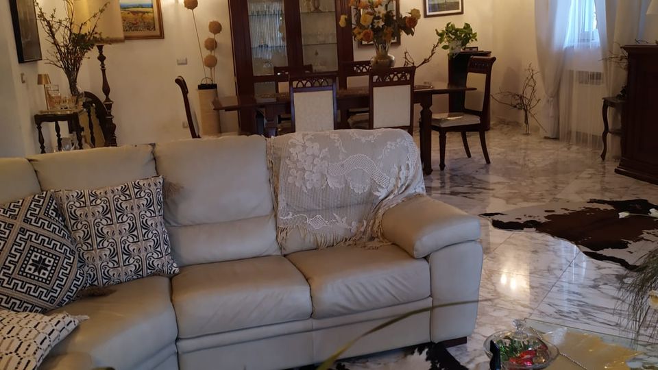 Soggiorno ampio con pavimento in marmo chiaro, divano ad angolo beige con cuscini decorativi e tappeti in stile pelle bovina. Sullo sfondo zona pranzo con tavolo e sedie, credenza in legno scuro e pareti con quadri e applique, accanto a una finestra con tende bianche.