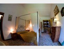Camera da letto in stile rustico con letto a baldacchino in ferro e tende bianche, copriletto dorato e comodini con lampade ai lati. Pavimento piastrellato decorato, pareti bianche con quadri e mobili in legno, con una finestra in fondo alla stanza.