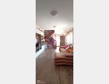 Soggiorno luminoso con pavimento in legno, grande divano a righe lungo la parete e parete attrezzata con TV e scaffali. Sul fondo si vede una scala interna in legno con ringhiera e una porta d’ingresso, con finestre laterali dotate di tende chiare.