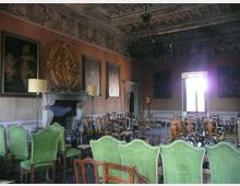 Sala storica con soffitto a cassettoni decorato e grande lampadario centrale, pareti con boiserie, dipinti e un camino in pietra. Numerose sedie d’epoca (anche con rivestimento verde) sono disposte in file, con una finestra luminosa sul fondo.