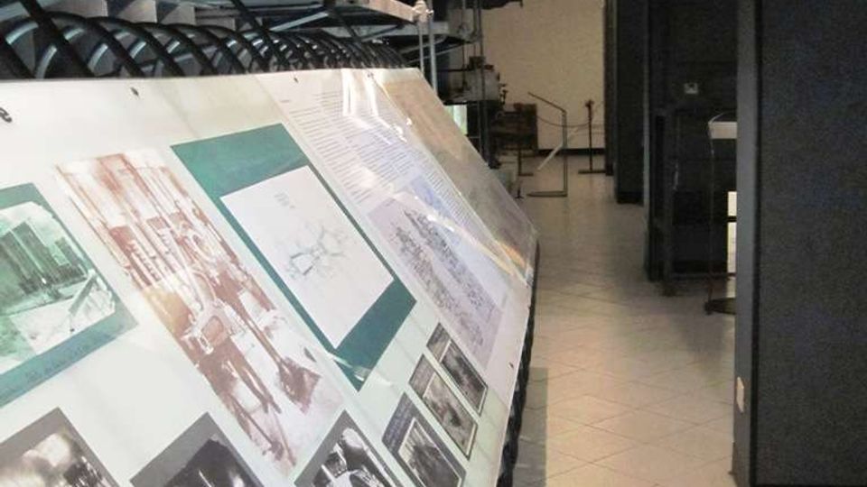 Corridoio tecnico industriale con pavimento piastrellato e grandi tubazioni e canalizzazioni a vista sul soffitto. In primo piano c’è un lungo pannello espositivo inclinato con fotografie e testi, affiancato da armadiature e macchinari scuri lungo il lato destro.