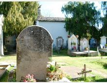 Piccolo cimitero all’aperto con lapidi e tombe in pietra, alcune con croci e fiori, su un prato. Sullo sfondo si vede un edificio bianco con tetto in tegole, circondato da alberi alti.