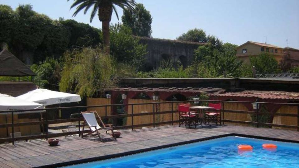 Piscina all’aperto con acqua azzurra e bordo in legno, circondata da una terrazza con ombrelloni, sedie a sdraio e tavolini. Sullo sfondo si vedono palme e vegetazione, tetti bassi e edifici sotto un cielo sereno.